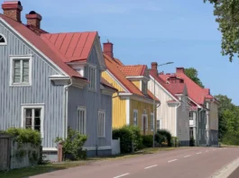 The image depicts a row of colorful, traditional wooden houses along a quiet street in Mariehamn, Åland Islands, Finland. The houses are painted in pastel shades of blue, yellow, and beige, each with a distinct, steeply sloped red metal roof. Architectural features include white-framed windows, decorative panels, and prominent chimneys. A narrow, empty road runs alongside the houses, bordered by a small patch of grass and various shrubs. Tall trees with lush green foliage are visible in the background under a clear blue sky, suggesting a bright, sunny day.