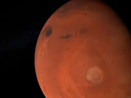 The image depicts a detailed view of the planet Mars against the backdrop of space. Mars is shown in its characteristic reddish-orange hue, dominating the right side of the image. The surface of the planet features various geological formations, including large, dark patches, potentially indicating volcanic regions or large basins. A prominent circular feature in the lower right quadrant appears to be a crater or volcanic dome, with surrounding textures suggesting dust and sediment distributions. The left side of the image fades into the darkness of space, speckled with faint, distant stars, emphasizing the isolation of the planet.
