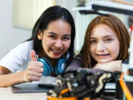 The image features two young girls sitting at a table, smiling at the camera. The girl on the left has long black hair and is wearing a white shirt. She has headphones resting around her neck and is giving a thumbs-up gesture. The girl on the right has shoulder-length brown hair and is leaning forward with her chin resting on her arms. Both girls are wearing braces. In the foreground, there is blurred electronic or robotic equipment, suggesting they might be engaged in a STEM activity. The setting appears to be a classroom or lab, with a bright window in the background.