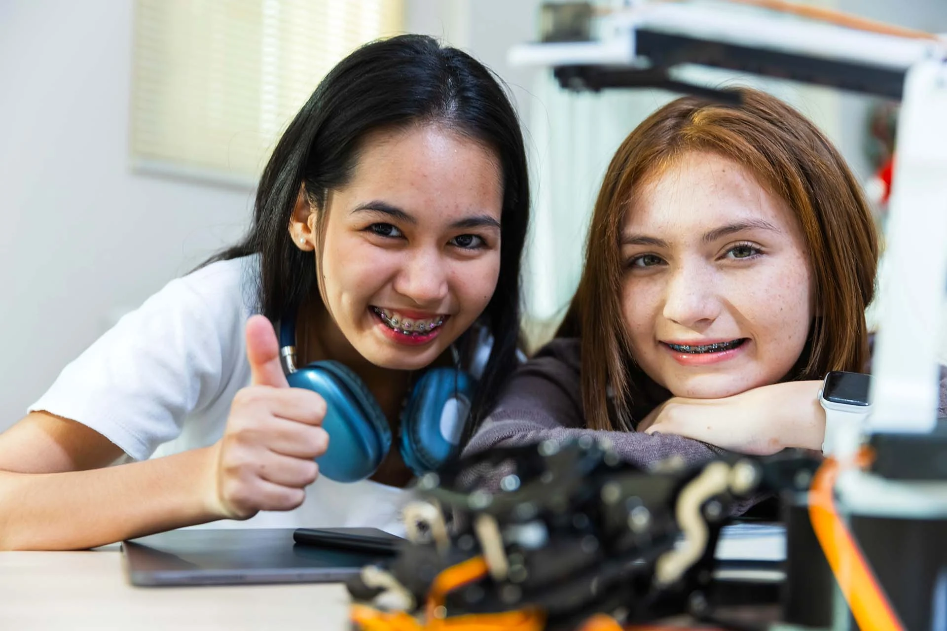 The image features two young girls sitting at a table, smiling at the camera. The girl on the left has long black hair and is wearing a white shirt. She has headphones resting around her neck and is giving a thumbs-up gesture. The girl on the right has shoulder-length brown hair and is leaning forward with her chin resting on her arms. Both girls are wearing braces. In the foreground, there is blurred electronic or robotic equipment, suggesting they might be engaged in a STEM activity. The setting appears to be a classroom or lab, with a bright window in the background.
