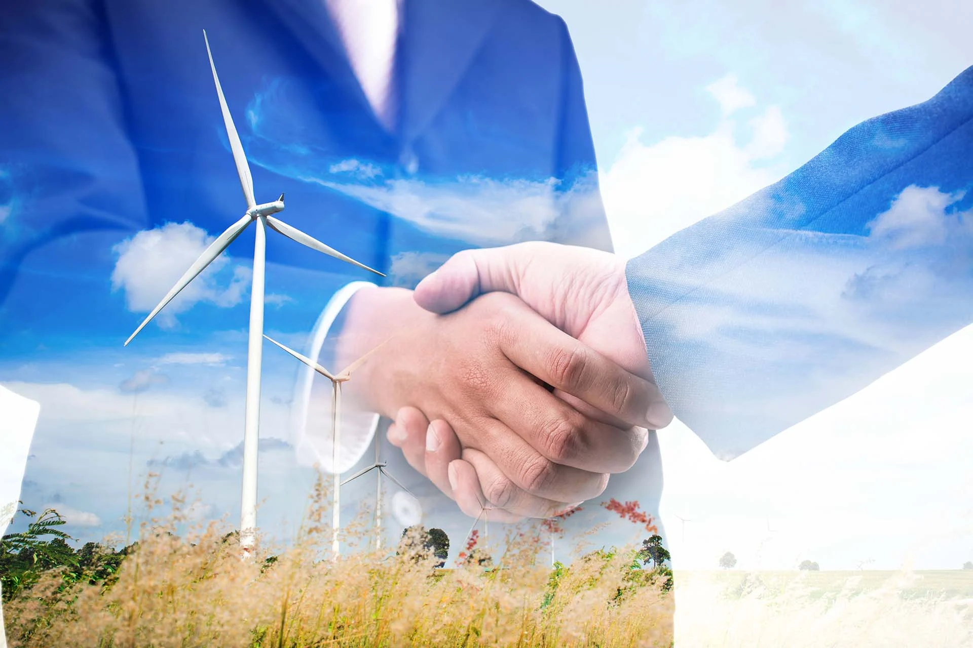 The image is a double-exposure composition featuring two primary elements: several wind turbines and a handshake between two individuals. In the foreground, two hands are clasped together in a firm handshake, symbolizing cooperation or agreement. The individuals are wearing blue suits, suggesting a business or professional context. In the background, a landscape with tall grass and multiple white wind turbines set against a bright blue sky with scattered clouds is visible. The turbines are tall with slender, three-bladed structures. The blending of the handshake with the environmental landscape hints at themes of sustainable business practices or agreements related to renewable energy.