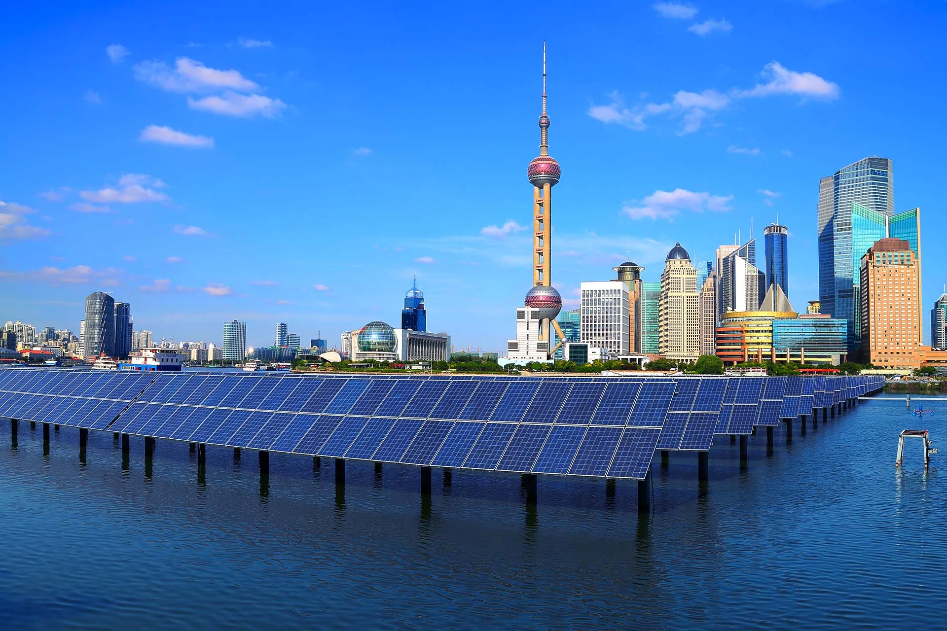 The image showcases a vibrant urban skyline featuring a prominent tower with a distinctive spherical design. In the foreground, there is an expanse of solar panels floating on a body of water. The solar panels are aligned in neat rows, creating a striking pattern against the water's surface. Behind the solar panels, modern skyscrapers with diverse architectural styles rise against a clear blue sky dotted with a few fluffy clouds. The cityscape includes buildings with varying heights and designs, featuring glass and steel structures.