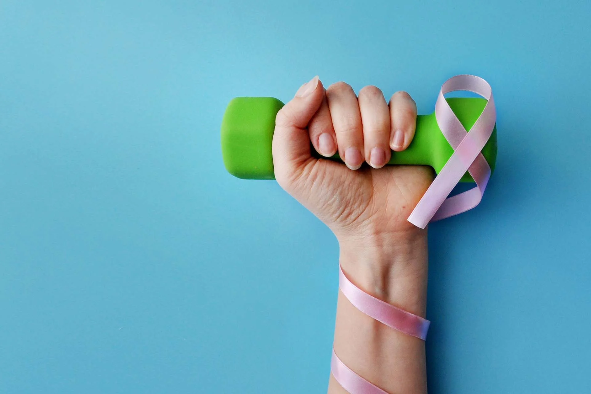 The image features a close-up of a hand gripping a small green dumbbell. A pink ribbon, commonly associated with breast cancer awareness, is wrapped around the dumbbell and hand, forming a loop over the fist. The background is a solid light blue, providing contrast to the green and pink colors. The skin tone of the hand is fair, and the nails are natural and unpainted.