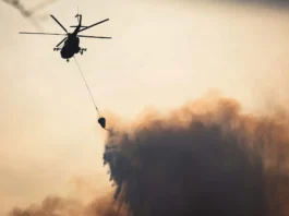 The image depicts a helicopter in silhouette against a hazy sky, engaged in aerial firefighting. The helicopter has a large rotor at the top and utility equipment attached to its underside. A long cable extends below the helicopter, suspending a bucket filled with water. The bottom of the image shows rising smoke, creating a dense, textured cloud of varying shades of brown and gray. The overall color palette is muted, with the smoke contrasting against the pale sky, adding dramatic effect to the scene.