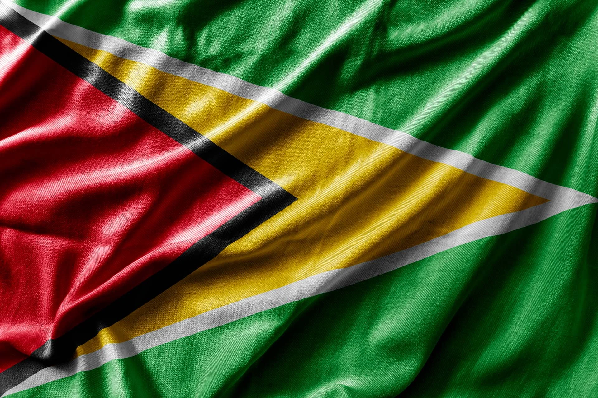 The image shows a detailed view of the flag of Guyana. The flag features a composition of geometric shapes in five colors: green, white, yellow, black, and red. The background consists of a green field. Along the hoist side, a red equilateral triangle, bordered by a narrow black stripe, points inward. A larger yellow triangle, edged in white, overlays the red triangle, also pointing inward. The fabric texture of the flag is visible, with folds adding depth and movement to the image.