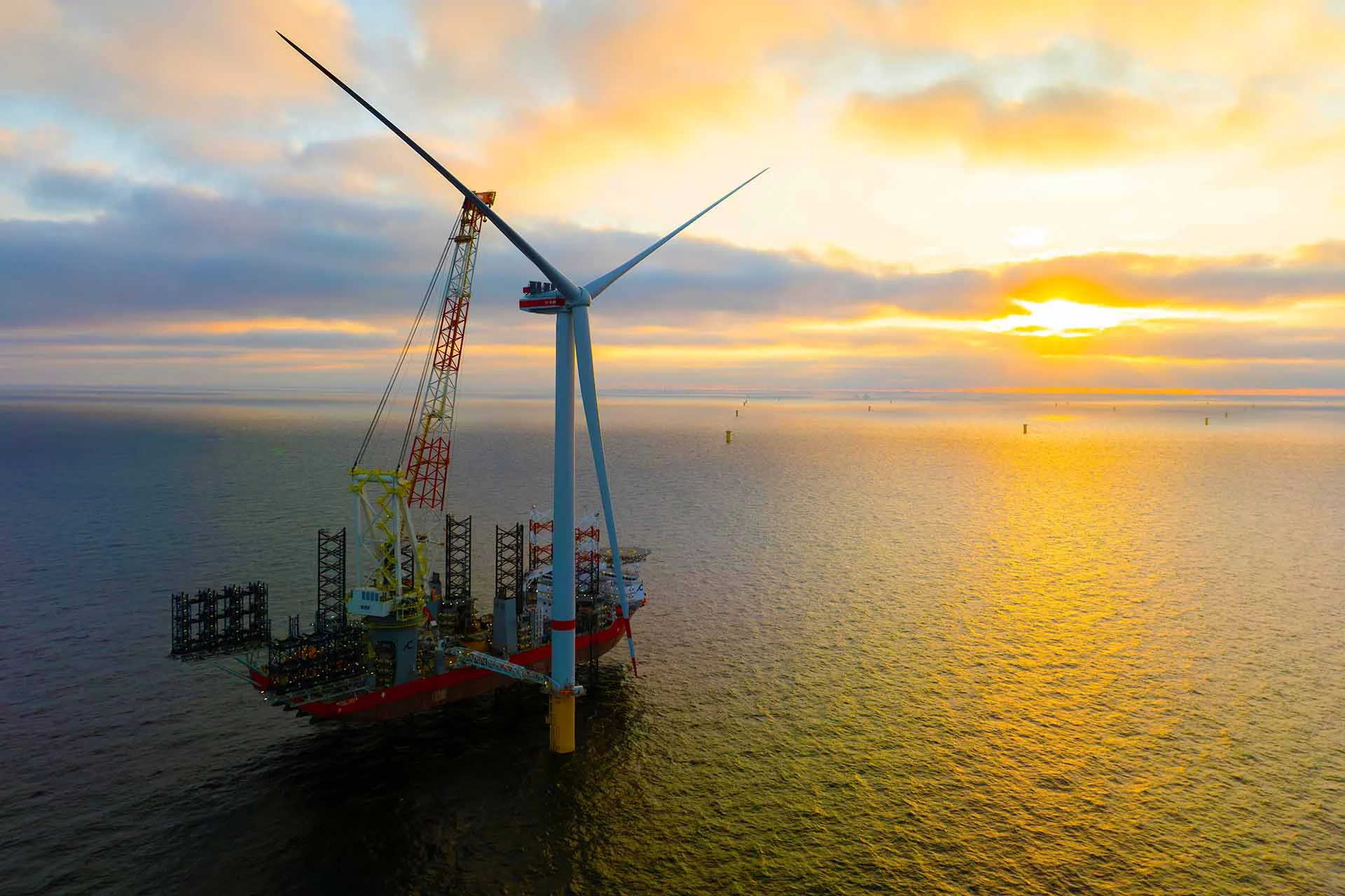 The image shows a large offshore wind turbine being installed at sea. The scene is set against a backdrop of a yellow and orange sunset over the ocean, with the sun partially obscured by clouds. The turbine itself is prominent in the foreground, featuring three long blades and a tall, sleek tower that rises from the water. A large vessel, equipped with cranes, is positioned alongside the turbine. This ship appears to be facilitating the installation process. The tranquil water reflects the warm tones of the sunset, creating a serene atmosphere.