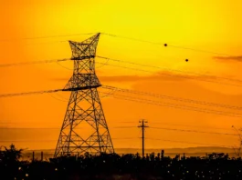 The image depicts a large electricity pylon set against a vibrant sunset sky in rural Kenya. The pylon, constructed from a lattice of metal beams, stands prominently on the left side of the composition. Several power lines extend horizontally across the frame, contributing a sense of structure and direction. The sky in the background is a rich, warm yellow transitioning to a deep orange, with soft, wispy clouds adding texture. Below the pylon, a silhouette of sparse vegetation is visible, enhancing the rural ambiance.