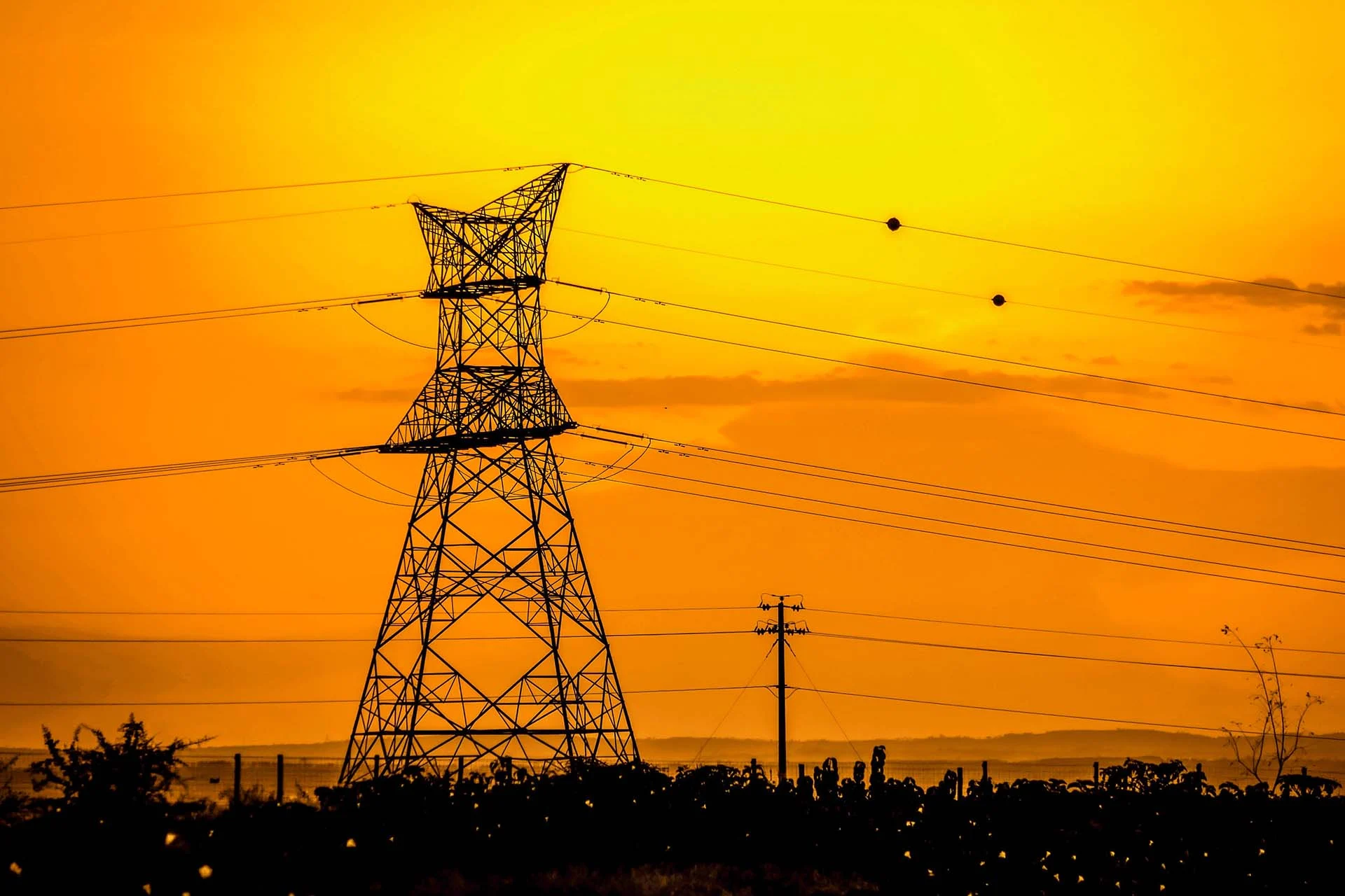 The image depicts a large electricity pylon set against a vibrant sunset sky in rural Kenya. The pylon, constructed from a lattice of metal beams, stands prominently on the left side of the composition. Several power lines extend horizontally across the frame, contributing a sense of structure and direction. The sky in the background is a rich, warm yellow transitioning to a deep orange, with soft, wispy clouds adding texture. Below the pylon, a silhouette of sparse vegetation is visible, enhancing the rural ambiance.