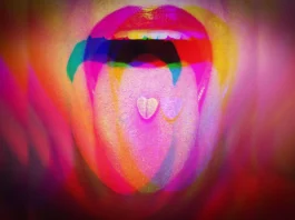 The image depicts a close-up of a person's mouth, slightly open with a visible tongue, set against a vibrant, multicolored background. The lips are painted a bright red, and on the tongue are two small, heart-shaped tablets. The overall color scheme features a blend of vivid pinks, purples, reds, greens, and yellows, creating an abstract and psychedelic effect. The overlapping colors and shapes give a dreamlike appearance to the composition.