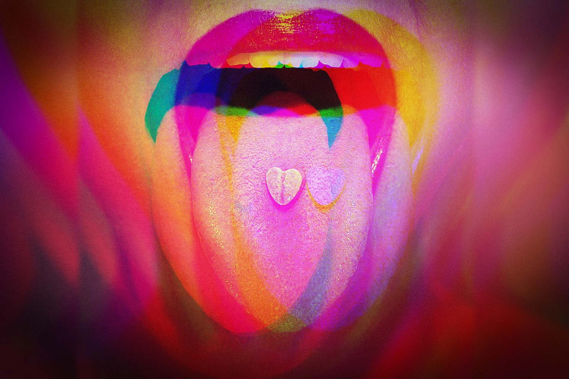 The image depicts a close-up of a person's mouth, slightly open with a visible tongue, set against a vibrant, multicolored background. The lips are painted a bright red, and on the tongue are two small, heart-shaped tablets. The overall color scheme features a blend of vivid pinks, purples, reds, greens, and yellows, creating an abstract and psychedelic effect. The overlapping colors and shapes give a dreamlike appearance to the composition.