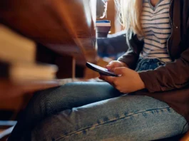 The image shows a close-up view of a person sitting at a wooden table. The focus is on their hands holding a smartphone, partially illuminated by sunlight. The individual is dressed in a striped shirt and blue jeans, with a dark jacket. The table surface is smooth and has a warm, rich tone. Positioned on the table are a stack of books, a pair of glasses, and a take-out coffee cup.