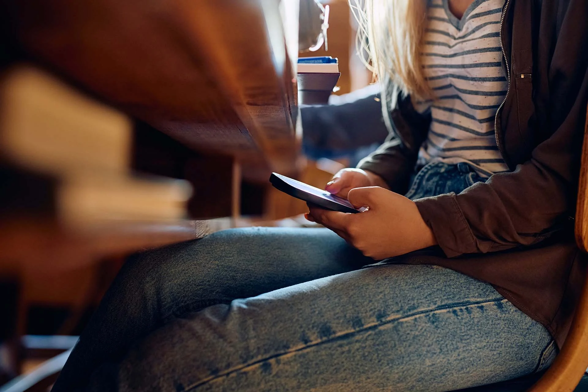 The image shows a close-up view of a person sitting at a wooden table. The focus is on their hands holding a smartphone, partially illuminated by sunlight. The individual is dressed in a striped shirt and blue jeans, with a dark jacket. The table surface is smooth and has a warm, rich tone. Positioned on the table are a stack of books, a pair of glasses, and a take-out coffee cup.