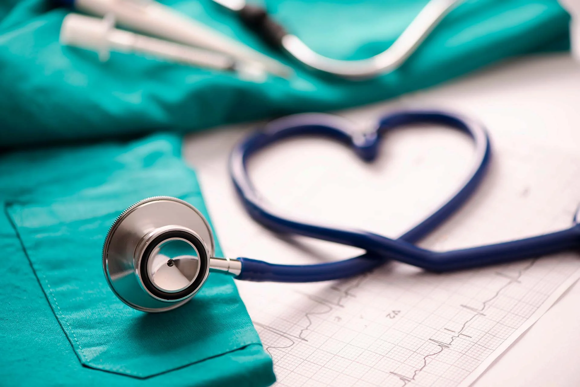 The image features a close-up view of a stethoscope placed prominently on a teal fabric, likely a medical scrub, with a visible pocket. The stethoscope has a metallic diaphragm and tubing that curves to form a heart shape on a white background. Beneath the tubing, there is a partially visible ECG (electrocardiogram) paper showing a graph pattern. The teal fabric stands in contrast to the white and blue tones of the stethoscope and paper, creating a clean and organized composition.