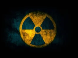 The image features a prominent radiation warning symbol. The symbol consists of a yellow circle with three equally spaced black triangles and a central black dot, forming the traditional radiation hazard icon. The background is a dark, textured surface that appears to be worn or aged, enhancing the starkness of the symbol. The yellow of the circle is vivid against the dark backdrop, emphasizing caution.