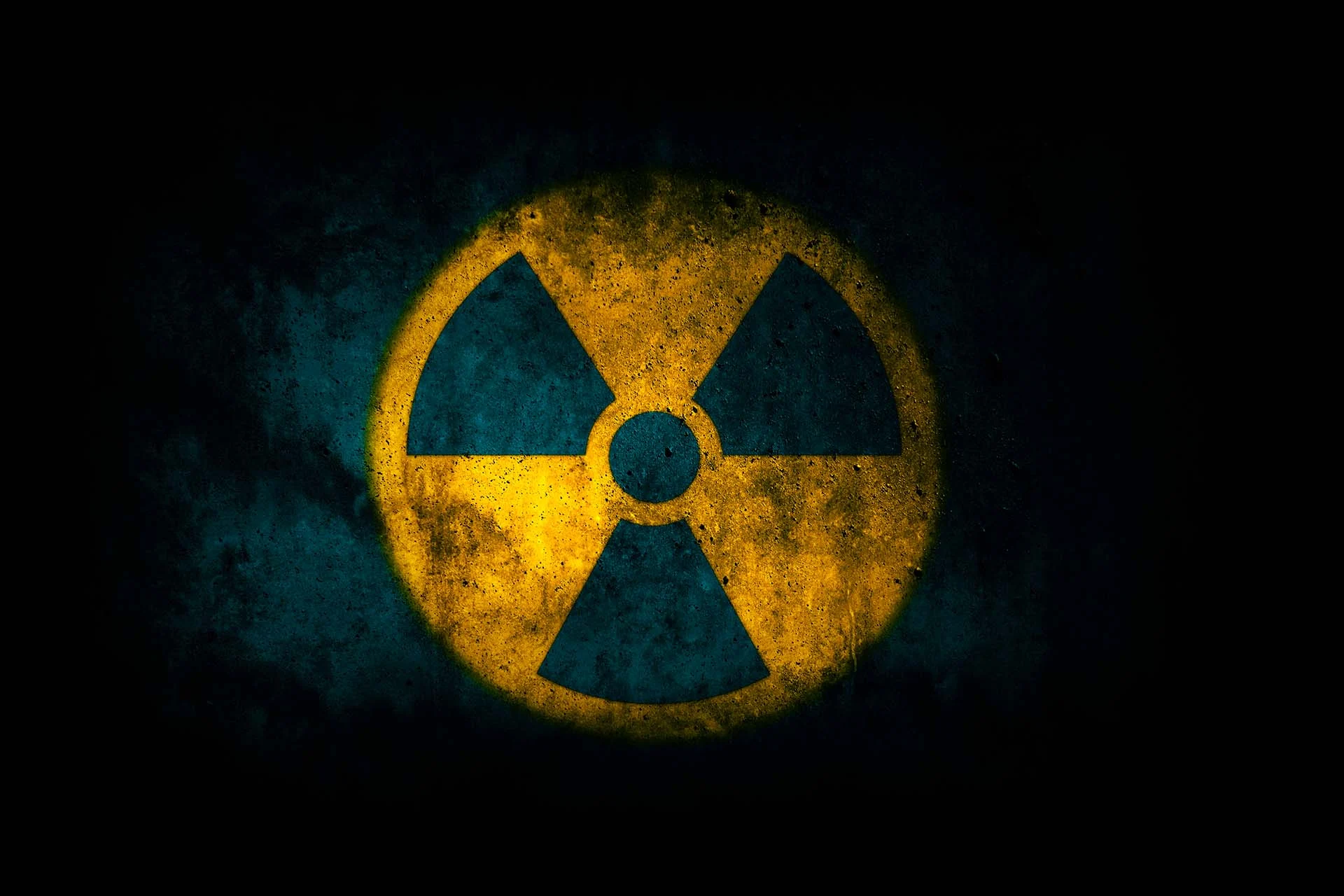 The image features a prominent radiation warning symbol. The symbol consists of a yellow circle with three equally spaced black triangles and a central black dot, forming the traditional radiation hazard icon. The background is a dark, textured surface that appears to be worn or aged, enhancing the starkness of the symbol. The yellow of the circle is vivid against the dark backdrop, emphasizing caution.