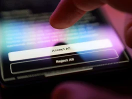 The image depicts a close-up view of a finger hovering over a smartphone screen. The screen displays two rectangular buttons with the options "Accept All" and "Reject All." The "Accept All" button is highlighted, suggesting interaction. The smartphone screen is illuminated, showing a blurred background with various colors, including blue and purple hues. The scene is presented in a dimly lit environment, creating a strong contrast between the screen's glow and the surrounding darkness.