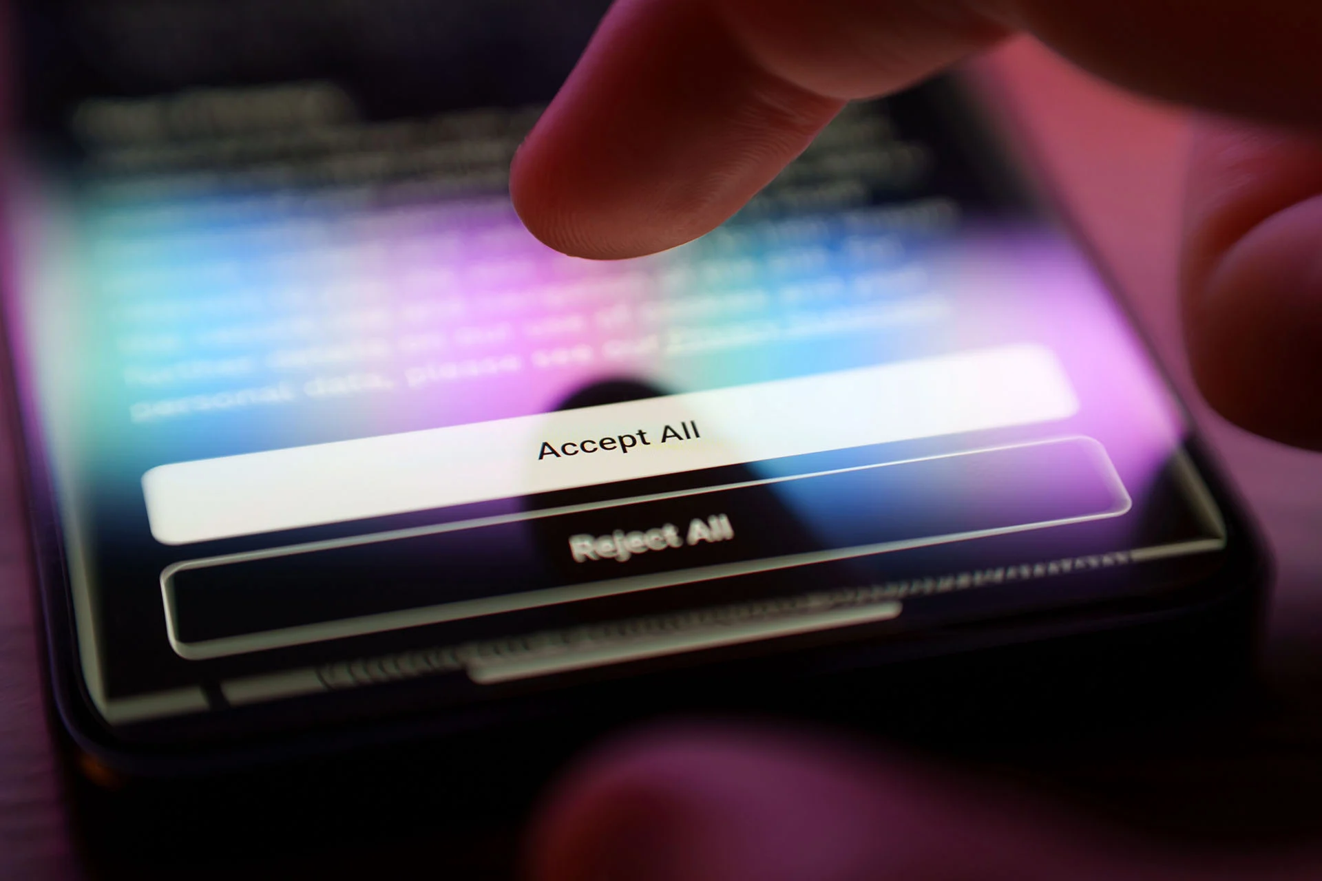 The image depicts a close-up view of a finger hovering over a smartphone screen. The screen displays two rectangular buttons with the options "Accept All" and "Reject All." The "Accept All" button is highlighted, suggesting interaction. The smartphone screen is illuminated, showing a blurred background with various colors, including blue and purple hues. The scene is presented in a dimly lit environment, creating a strong contrast between the screen's glow and the surrounding darkness.