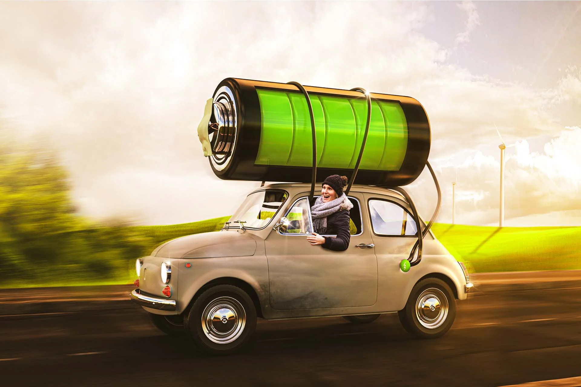 The image depicts a whimsical scene of a small vintage car driving on a road in a countryside setting. The car is noticeable for the large, green battery mounted on its roof, with thick cables connecting it to the vehicle, suggesting the idea of an electric car. The battery is oversized compared to the car, with a bright green color indicating full charge. A person dressed in winter clothes, including a black hat and scarf, is joyfully leaning out of the car’s window. The background features blurry green fields and several wind turbines scattered across the landscape, under a cloudy sky tinged with shades of orange and yellow, implying a sunset or sunrise.