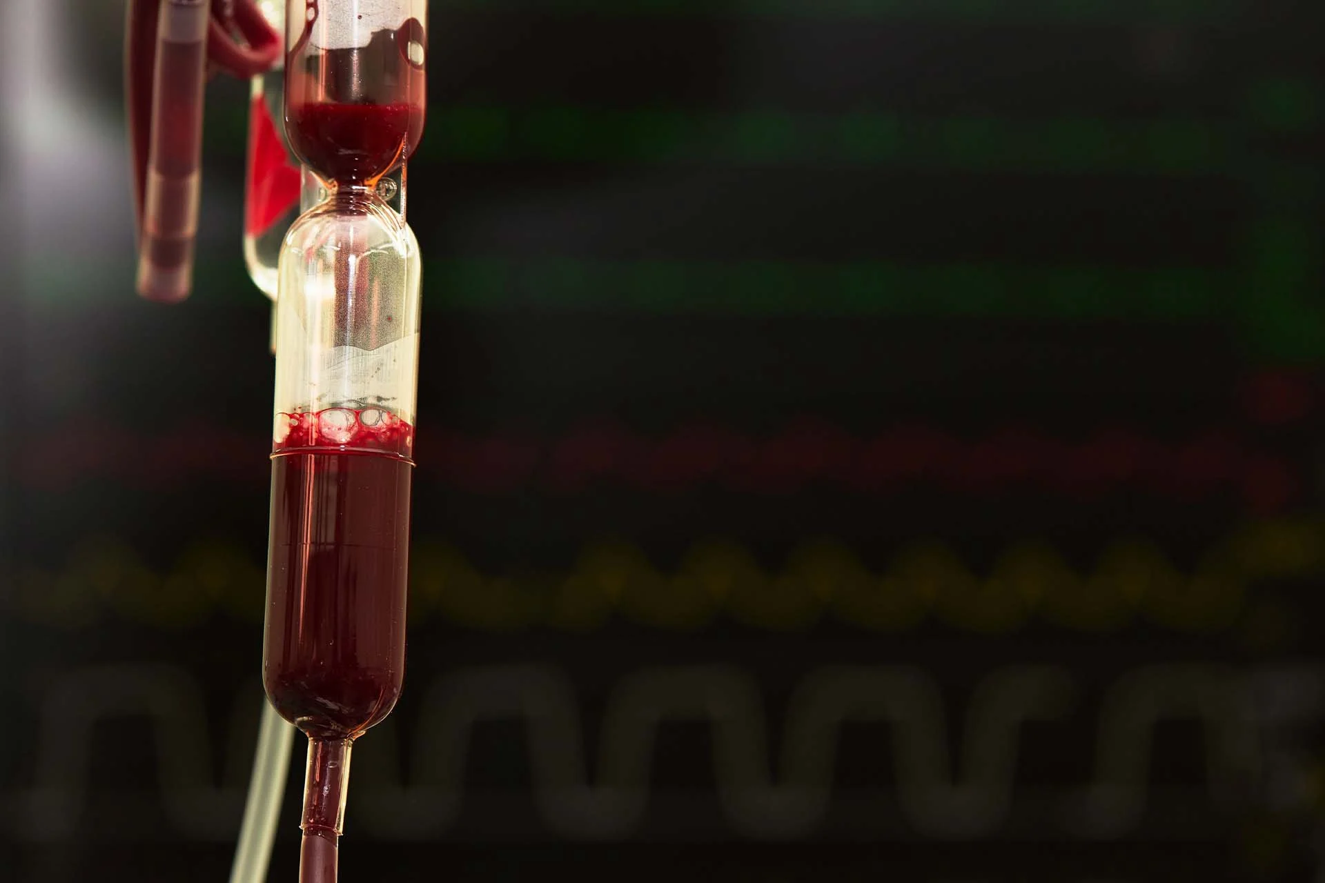 The image shows a close-up view of a blood transfusion chamber. The chamber contains a clear liquid mixed with red blood, which creates a gradient of colors from pale red to rich crimson. Small air bubbles are visible near the surface of the liquid. The transfusion chamber is vertically oriented and connected to tubes, with some appearing above and others below the chamber. The background displays blurred dark monitors with green, red, and yellow waveform patterns, typical of hospital equipment.