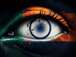 The image portrays a close-up of a human eye with the iris transformed into the Ashoka Chakra, a 24-spoke wheel from the Indian flag. Surrounding the eye, the eyelid and surrounding skin are painted with the colors of the Indian flag: saffron at the top, white in the middle, and green at the bottom. The vibrant colors are slightly blended, giving a painted effect. The eye's lashes are clearly defined and sharply contrasted against the vivid colors.