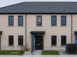 The world’s first 3D-printed concrete social housing project stands in Grange Close, Dundalk, County Louth, Ireland, a terrace of three identical, modern 3-bedroom homes. The image shows a sleek, two-story structure with a light beige exterior and dark gray sloped roofs. Each unit features large black-framed windows, two on each floor, and the central home includes a black front door with a small rectangular window and a minimalist flat awning. Gray stone paths lead from the sidewalk to each entrance, bordered by low black railings. The small front yards are landscaped with green grass and tall ornamental planters, reflecting a clean, contemporary design.