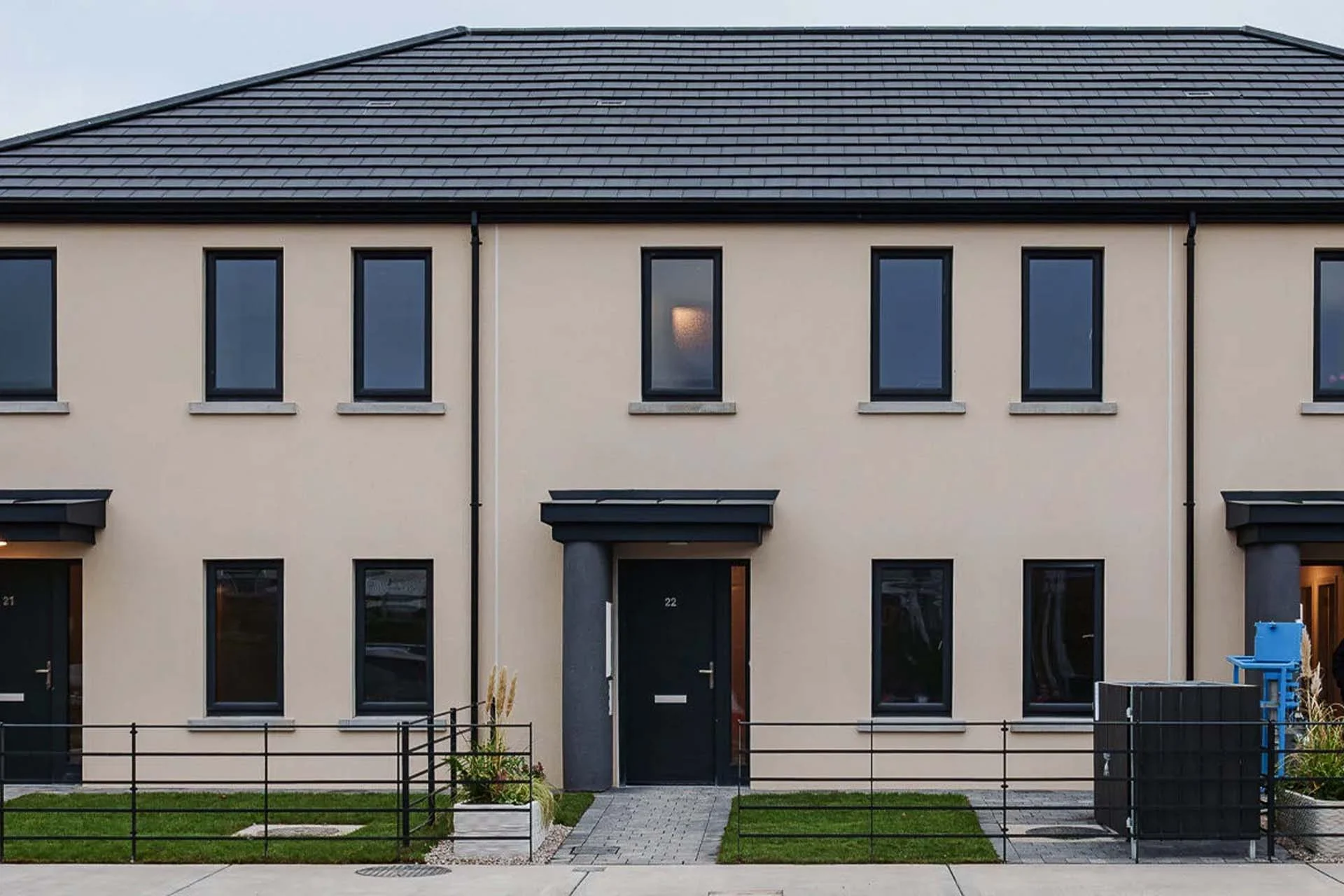 The world’s first 3D-printed concrete social housing project stands in Grange Close, Dundalk, County Louth, Ireland, a terrace of three identical, modern 3-bedroom homes. The image shows a sleek, two-story structure with a light beige exterior and dark gray sloped roofs. Each unit features large black-framed windows, two on each floor, and the central home includes a black front door with a small rectangular window and a minimalist flat awning. Gray stone paths lead from the sidewalk to each entrance, bordered by low black railings. The small front yards are landscaped with green grass and tall ornamental planters, reflecting a clean, contemporary design.