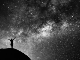 The image is a black and white photograph depicting a silhouette of a person standing on a hill or rocky outcrop with arms outstretched against a backdrop of a star-filled night sky. The vast expanse of the sky is densely populated with stars of varying brightness, creating a shimmering effect across the image. In the center, there is a denser cluster of stars, possibly indicating the Milky Way galaxy. The contrast between the dark silhouette of the person and the bright, starry sky adds a dramatic and awe-inspiring effect to the scene.