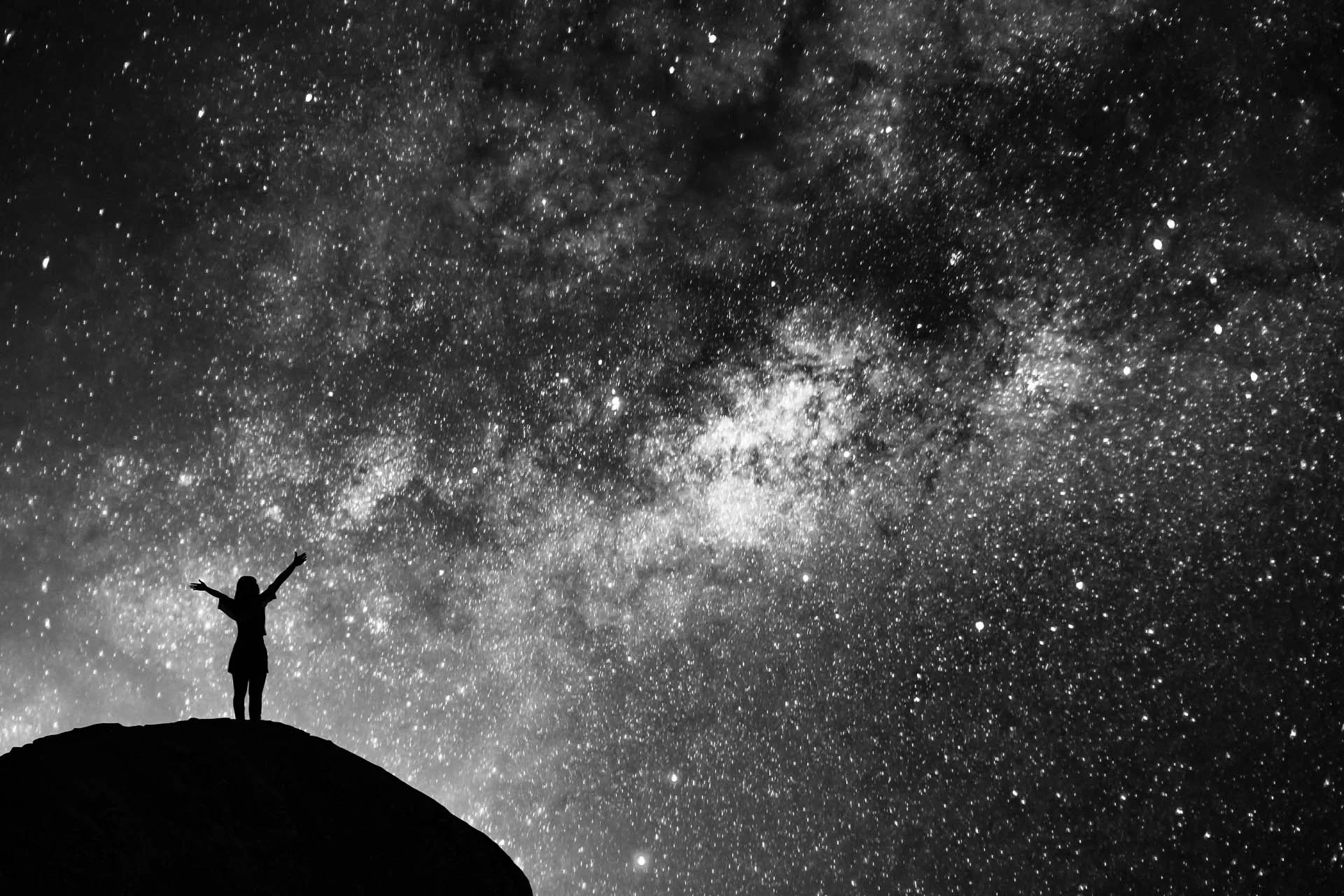The image is a black and white photograph depicting a silhouette of a person standing on a hill or rocky outcrop with arms outstretched against a backdrop of a star-filled night sky. The vast expanse of the sky is densely populated with stars of varying brightness, creating a shimmering effect across the image. In the center, there is a denser cluster of stars, possibly indicating the Milky Way galaxy. The contrast between the dark silhouette of the person and the bright, starry sky adds a dramatic and awe-inspiring effect to the scene.