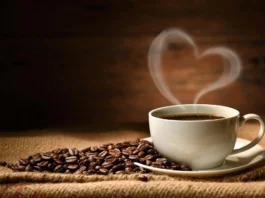 The image features a white ceramic cup filled with dark coffee, resting on a matching saucer. The cup is positioned to the right on a textured burlap surface. A pile of shiny, brown coffee beans is scattered attractively around the cup. Rising from the hot coffee is steam in the shape of a heart, adding a warm and inviting touch to the composition. The background is softly blurred, emphasizing the rustic wooden texture, which enhances the cozy and intimate atmosphere of the scene.