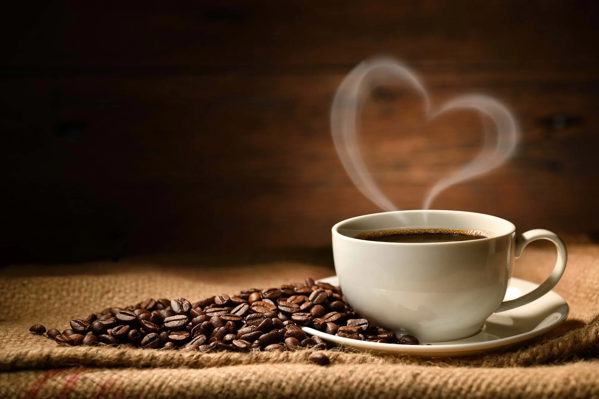 The image features a white ceramic cup filled with dark coffee, resting on a matching saucer. The cup is positioned to the right on a textured burlap surface. A pile of shiny, brown coffee beans is scattered attractively around the cup. Rising from the hot coffee is steam in the shape of a heart, adding a warm and inviting touch to the composition. The background is softly blurred, emphasizing the rustic wooden texture, which enhances the cozy and intimate atmosphere of the scene.