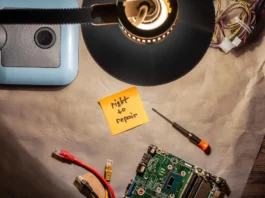 The image shows a dimly lit workspace with various electronics and tools displayed on a brown paper surface. A lamp with a circular shade illuminates the area from above, casting a soft light. To the left, there is a blue and gray device, partially visible under the lamp's shade. A prominent yellow sticky note near the center reads "right to repair." Surrounding it, wires with colored casings, an electronic circuit board, a small flathead screwdriver with an orange handle, and two network cables, one red and another yellow, are scattered around.